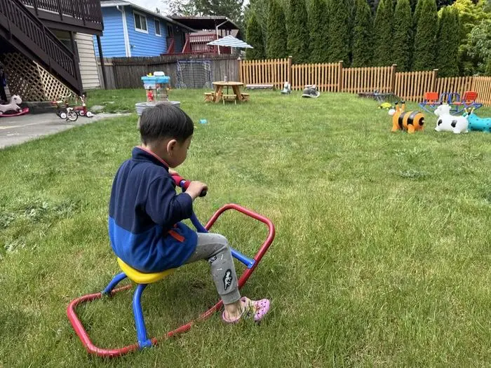 
Child sitting on outdoor play equipment: A toddler happily seated on a low, colorful swing in a safe daycare playground surrounded by open outdoor space.